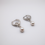 Load image into Gallery viewer, A pair of silver huggie hoop earrings with a small chain link and a single white pearl dangle, shown on a plain light gray background.
