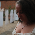 Load image into Gallery viewer, A woman in a white lace-trimmed dress wearing a silver necklace with a small white pearl pendant, photographed outdoors with a blurred brick wall and white garments in the background.
