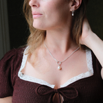 Load image into Gallery viewer, A close-up lifestyle shot of a woman wearing a delicate silver necklace with a single white pearl pendant and a matching silver drop earring. She is wearing a dark brown smocked blouse with white lace trim.
