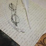 Load image into Gallery viewer, A silver wire-frame eyeglasses necklace with a single white pearl pendant, styled on top of vintage handwritten paper alongside a white quill pen.
