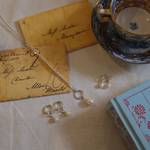 Load image into Gallery viewer, A silver-toned necklace and matching drop earrings with small pearl-like beads are arranged on a cream fabric surface with vintage paper notes, a blue-and-gold teacup and saucer, and a light blue book.
