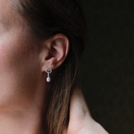 Load image into Gallery viewer, A close-up side profile of a woman wearing a delicate silver hoop earring with a dangling white pearl, set against a dark, shadowed background.
