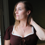 Load image into Gallery viewer, A woman with light brown hair wearing a dark brown textured top with a white lace-trimmed neckline and a pearl pendant necklace, looking off-camera in a dimly lit interior.

