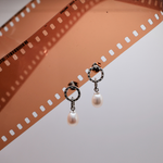 Load image into Gallery viewer, A pair of silver-toned drop earrings with circular ring details and dangling white pearl beads, displayed on a film strip against a light gray background.
